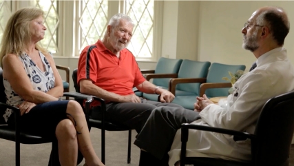 Video play button with an image of someone living with Parkinson’s, his care partner, and doctor